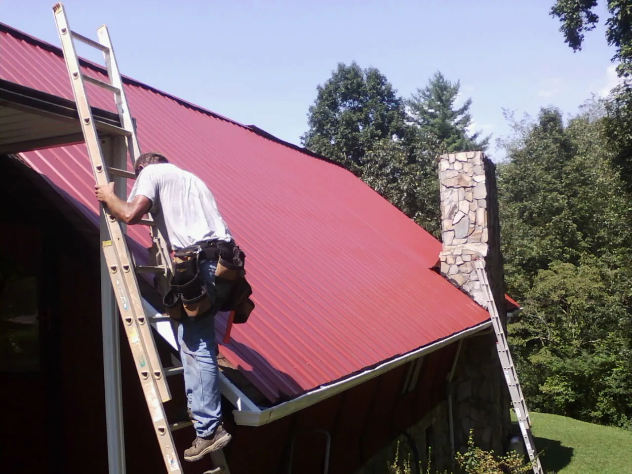Roofer on a ladder beside a red metal roof and stone chimney — Soffit & Fascia Repair by Roofing Brothers in Menan